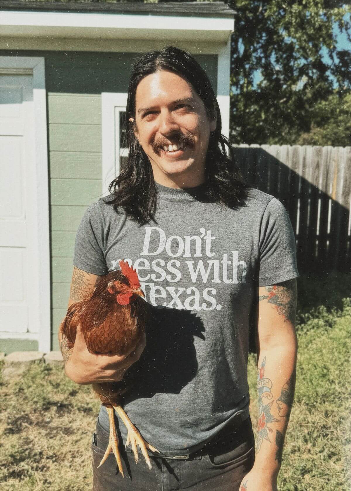 Eric Solis standing in yard, holding a rooster, wearing a shirt that says, "Don't mess with Texas."