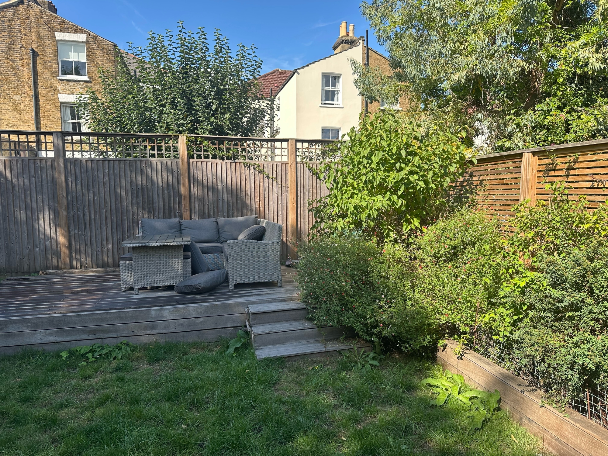 Before photo of South London family garden showing raised timber deck with rattan furniture, patchy lawn and mature shrubs