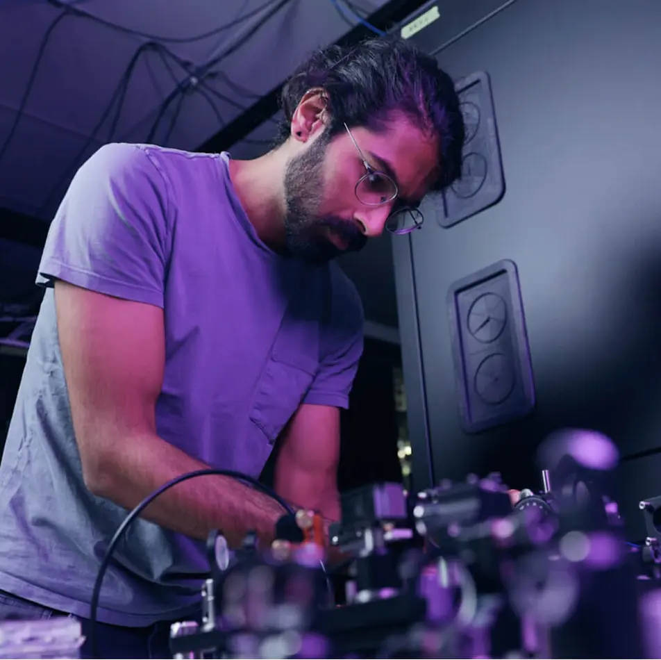 Man wearing glasses and a purple shirt working with scientific equipment in a lab illuminated by purple lighting.