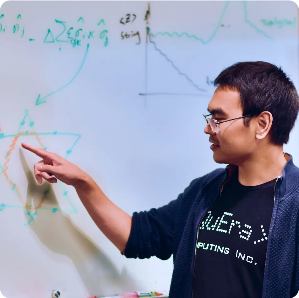 Man wearing glasses and a black t-shirt pointing at a whiteboard with mathematical equations and graphs.