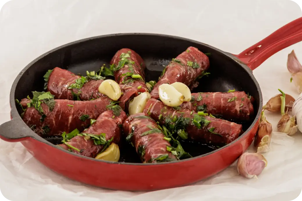 מחבת אדומה עם גלילות בשר עטופות עשבי תיבול ושיני שום, מוקפצות בשמן.