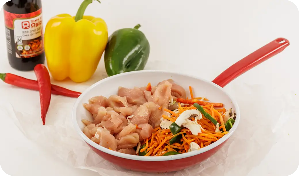 מחבת אדומה עם חתיכות עוף גולמיות וירקות כולל גזר, פטריות ופלפל ירוק לצד פלפל צהוב, פלפל ירוק, שני פלפלים אדומים ובקבוק רוטב