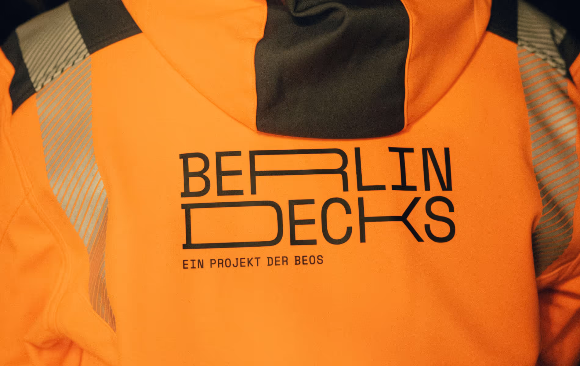 A close-up photo of the back of an orange jacket featuring the Brand Identity of "BERLIN DECKS"