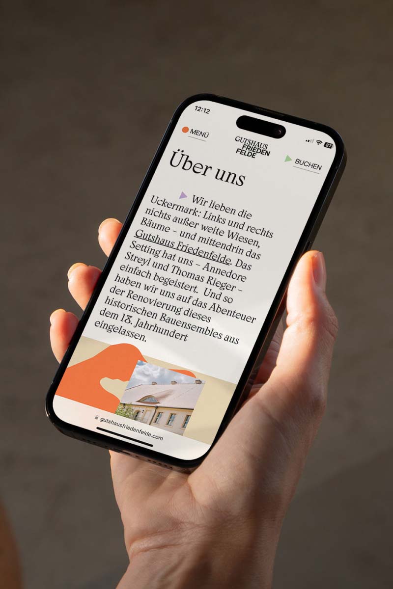 A hand holding a phone showing the website design of the Gutshause Friedenfelde about us page