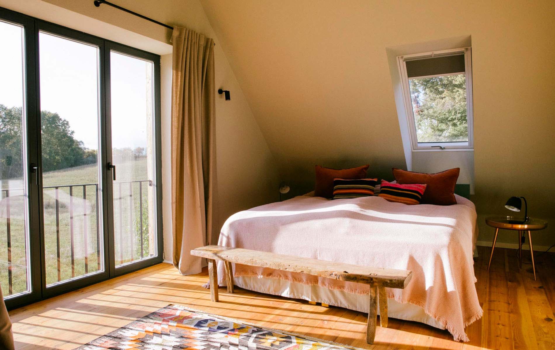 Cosy bedroom with a big bed that uses white sheets and colorful pillows. In front of the bed a small wooden bench is placed. The room looks out to green fields.