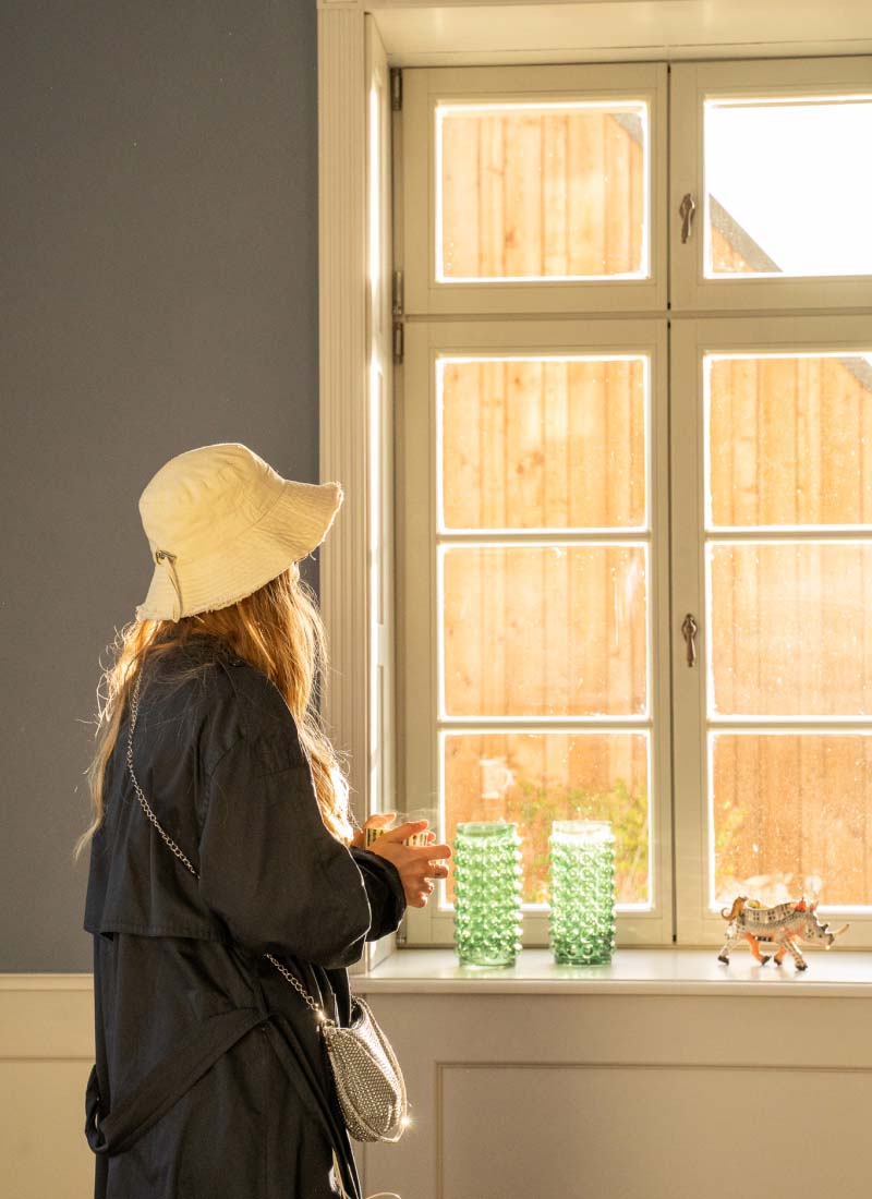 A person wearing a sun hat holding a hot drink in their hands standing in a room looking out of the windows