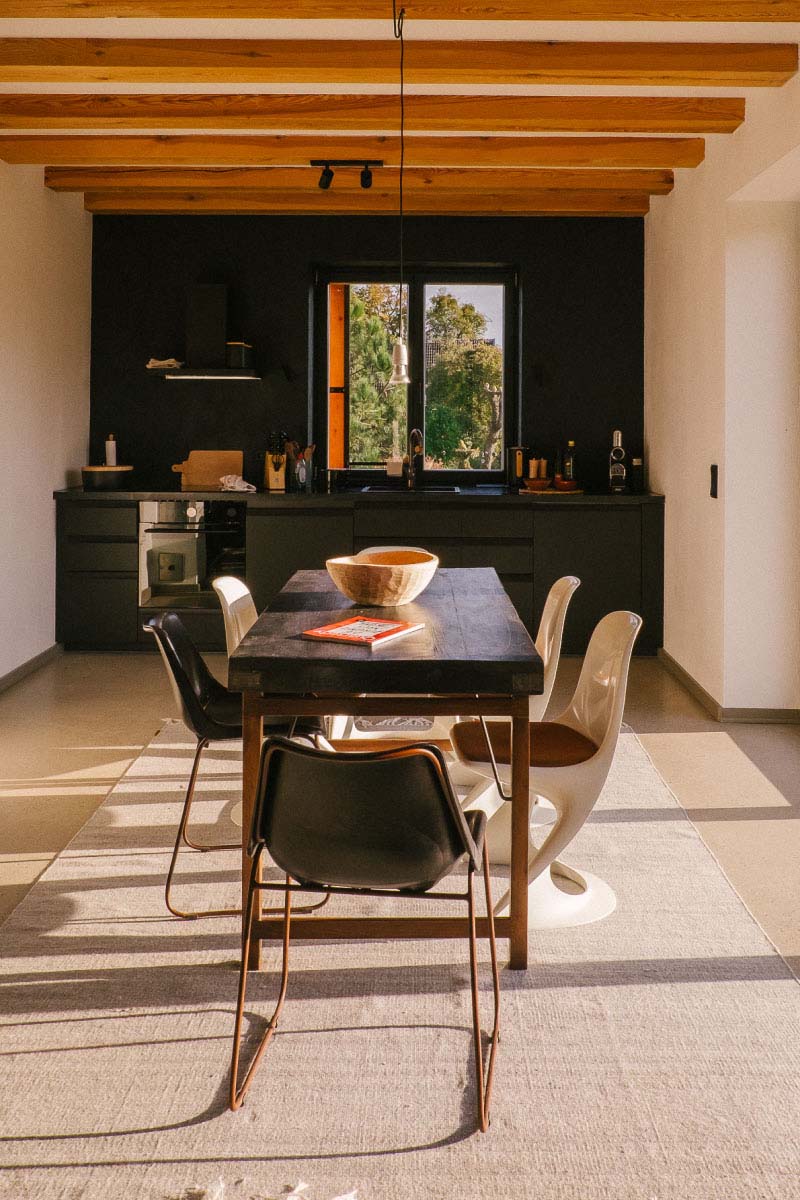 One of the big eating tables with many chairs placed around it with the kitchen in the back of the sunlight room
