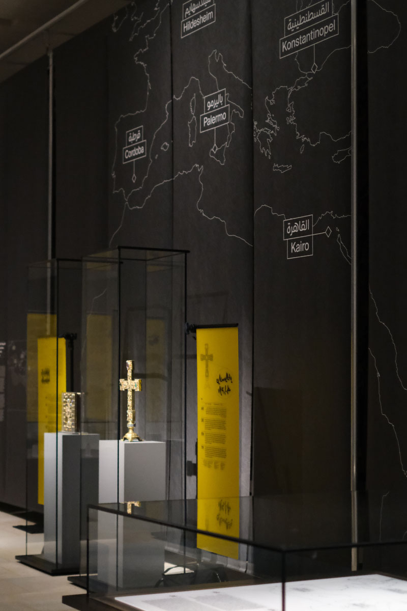 Photo with several exhibits in a museum with Yellow information boards. On a wall is a european map with Kairo, Palermo, Cordoba, Konstantinopel and Hildesheim.