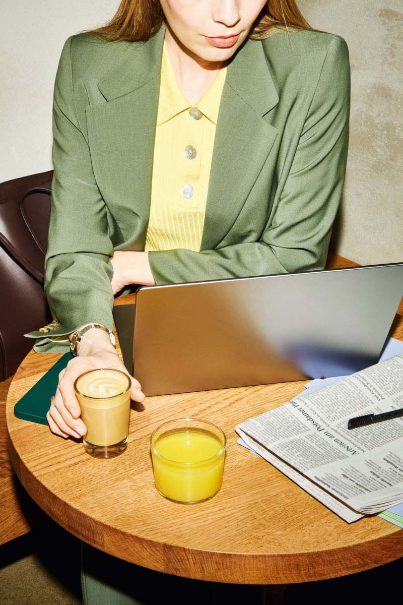 Photo of a person in a green blazer sitting at a table with newspapers and a laptop drinking coffee and orange juice, the shot is part of the marketing campaign