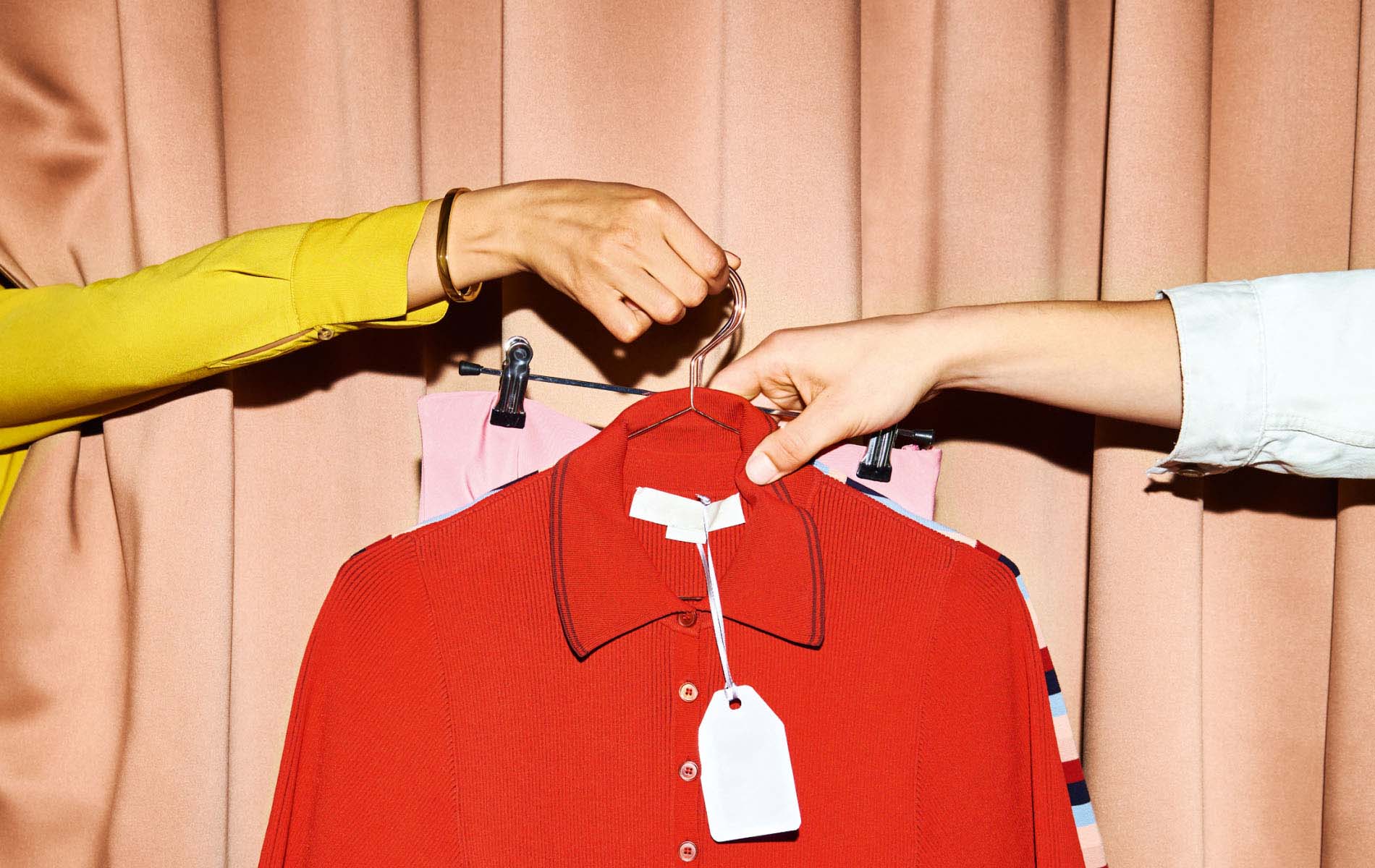 Two arms holding up several pieces of clothing on hangers in front of a peach colored curtain, the shot is part of the marketing campaign