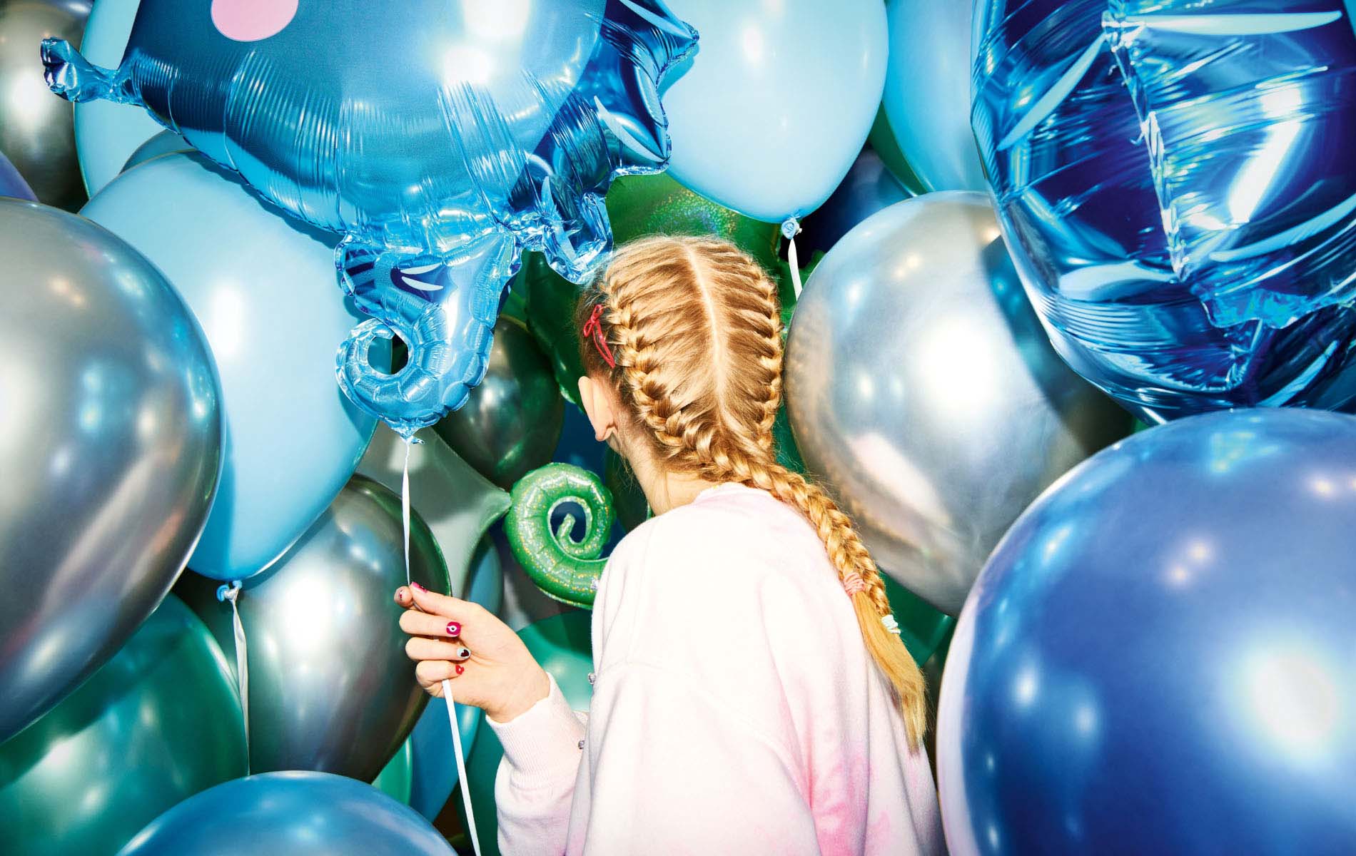 A blonde girl with a pink sweater is surrounded in blue, silver and green balloons, the shot is part of the marketing campaign