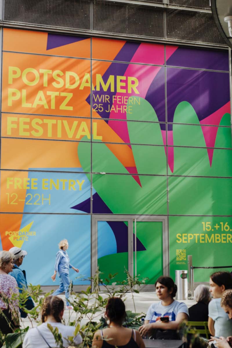 A group of people sitting under a large wall graphic that reads "Potsdamer Platz Festival - Wir feiern 25 Jahre" on a background with shapes in four different colors, showing the brand experience