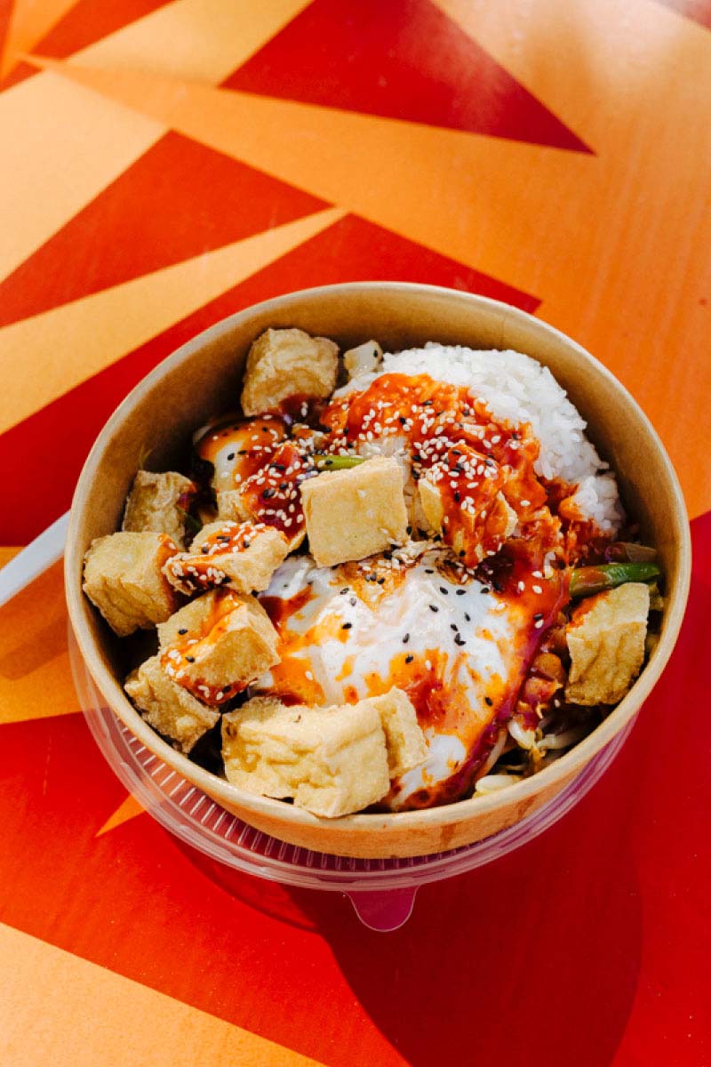 A bowl of food with Tofu and rice, topped with hot sauce on a red table.