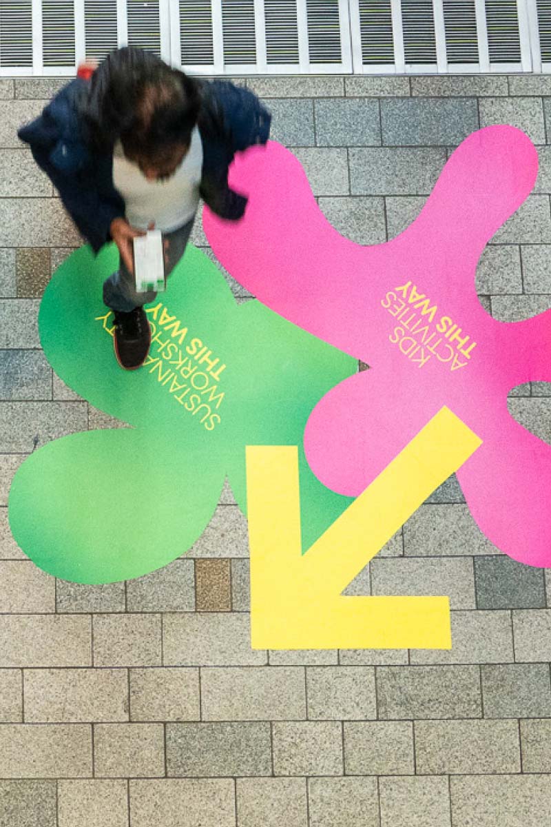Person walking over the on-ground wayfinding depicting two shapes with yellow text and a bold yellow directional arrow, as part of the brand identity design