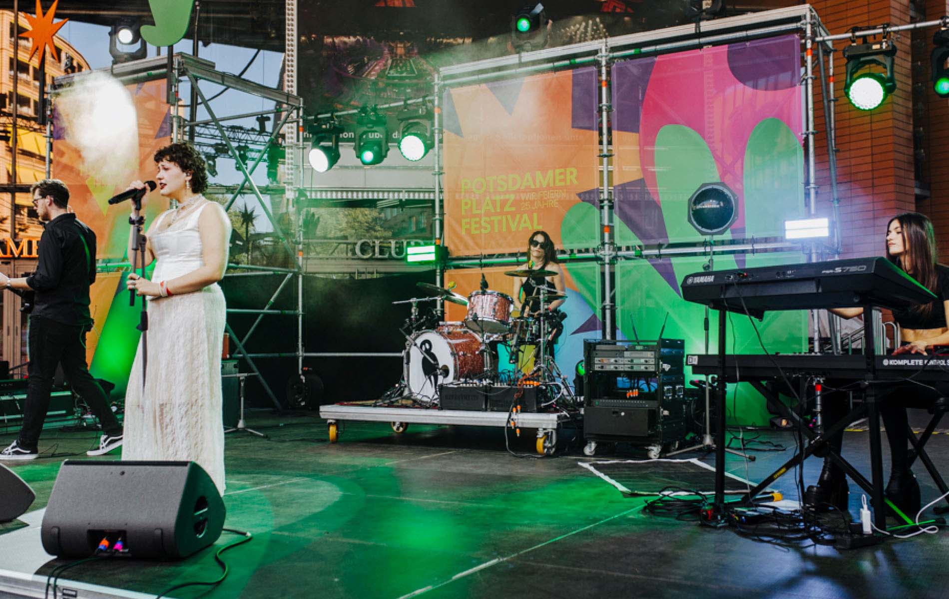 A singer in a white dress on stage with a band and the Potsdamer Platz design in the back aswell as several cut out shapes on the stage, showing the brand experience