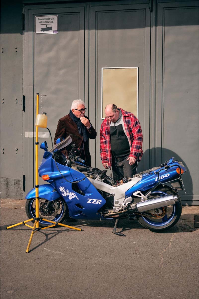 Two old men in front of a steal door standing behind a motorcycle and inspecting it