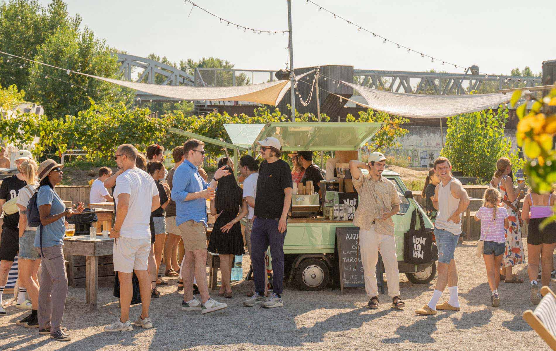 A crowd of people gathered around an italian ape selling coffee in a park 