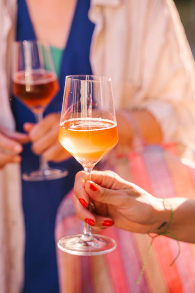 A hand holding a glass of orange wine with another blurry hand and glass of wine in the background