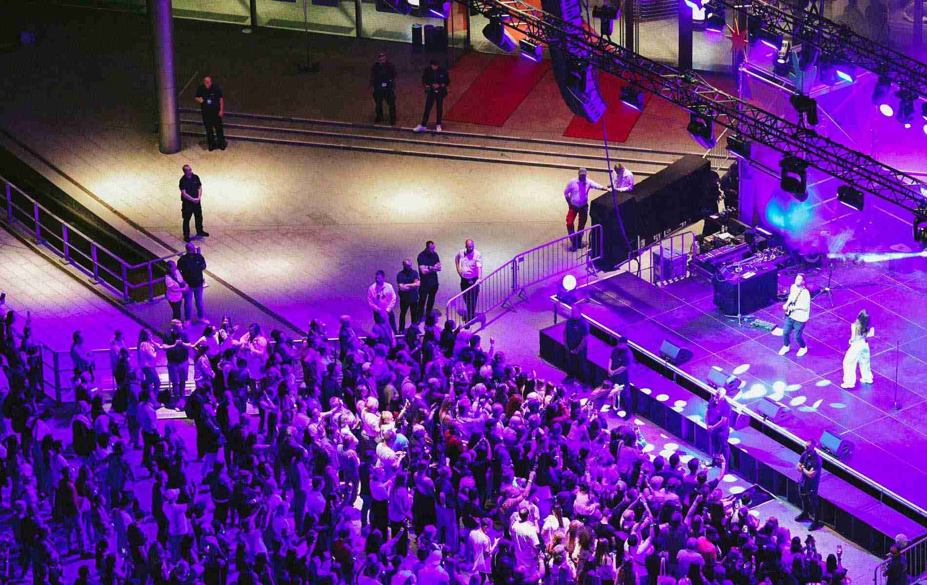 A concert stage with a large crowd bathed in a purple light from a bird's eye view, showing the event experience