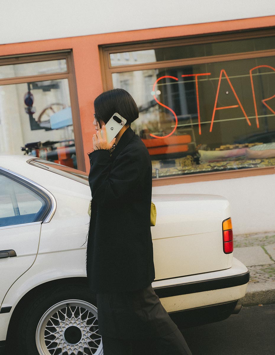A woman walking beside a vintage car while talking on a Google Pixel phone, created by a Creative Agency in Berlin producing Social Media Content, Photography for Brands, and Branding visuals.