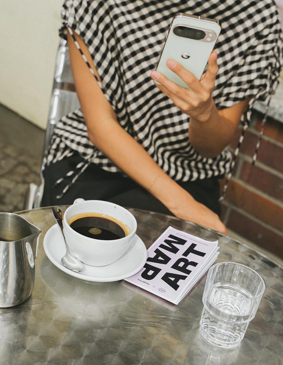 A woman sitting at an outdoor café holding a Google Pixel phone, with coffee and an art map on the table, shot by a Creative Agency for Content Marketing, Social Media Content, and Editorial Photography.