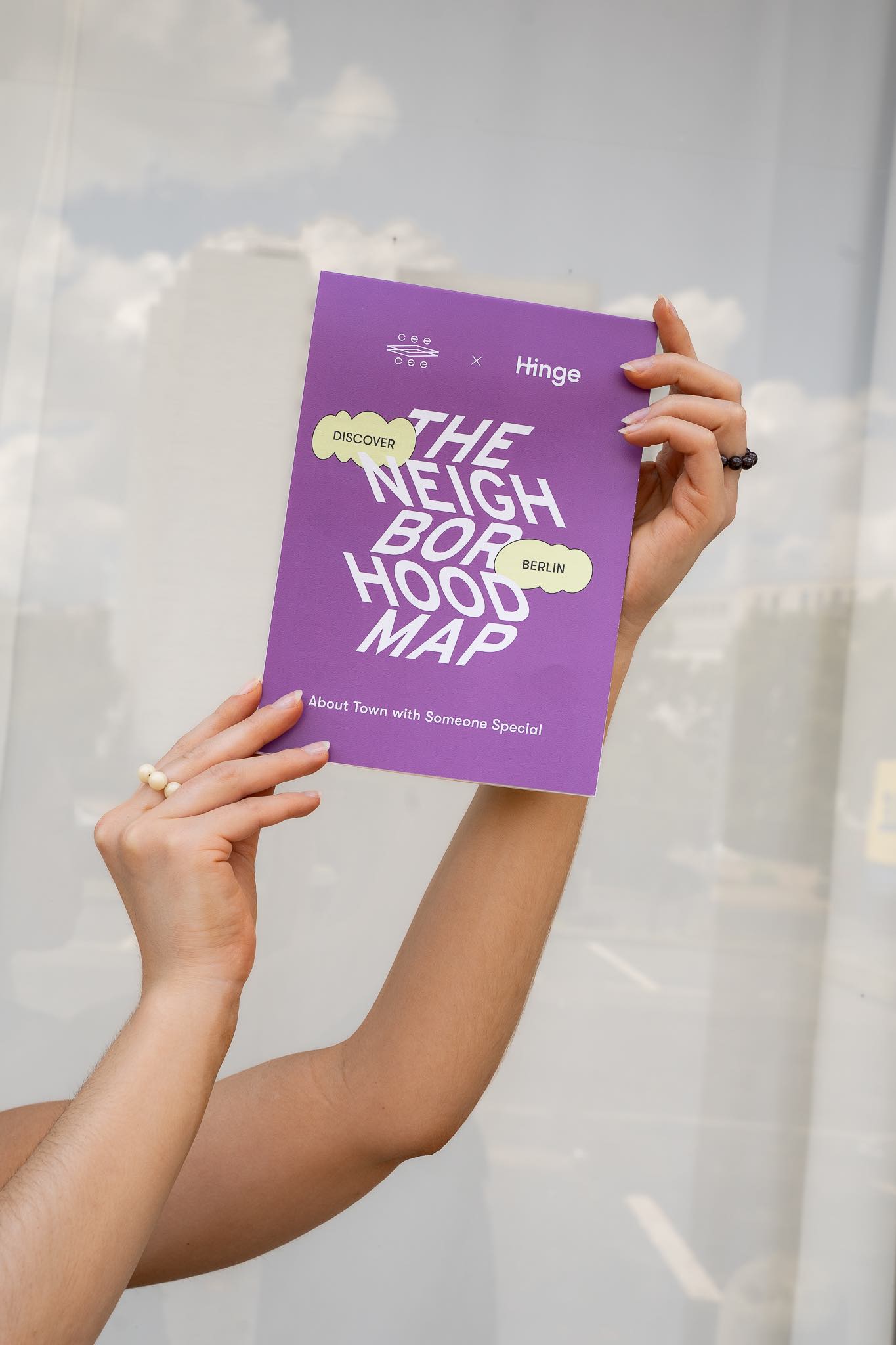 A person holds a purple “The Neighborhood Map” brochure up toward the sky in front of a reflective window façade.