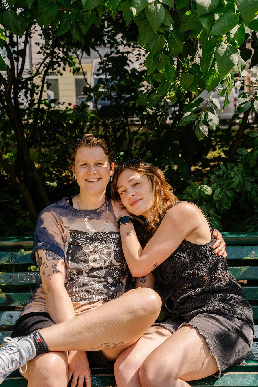 A couple sits close together on a park bench, smiling and leaning into each other, surrounded by lush green leaves.