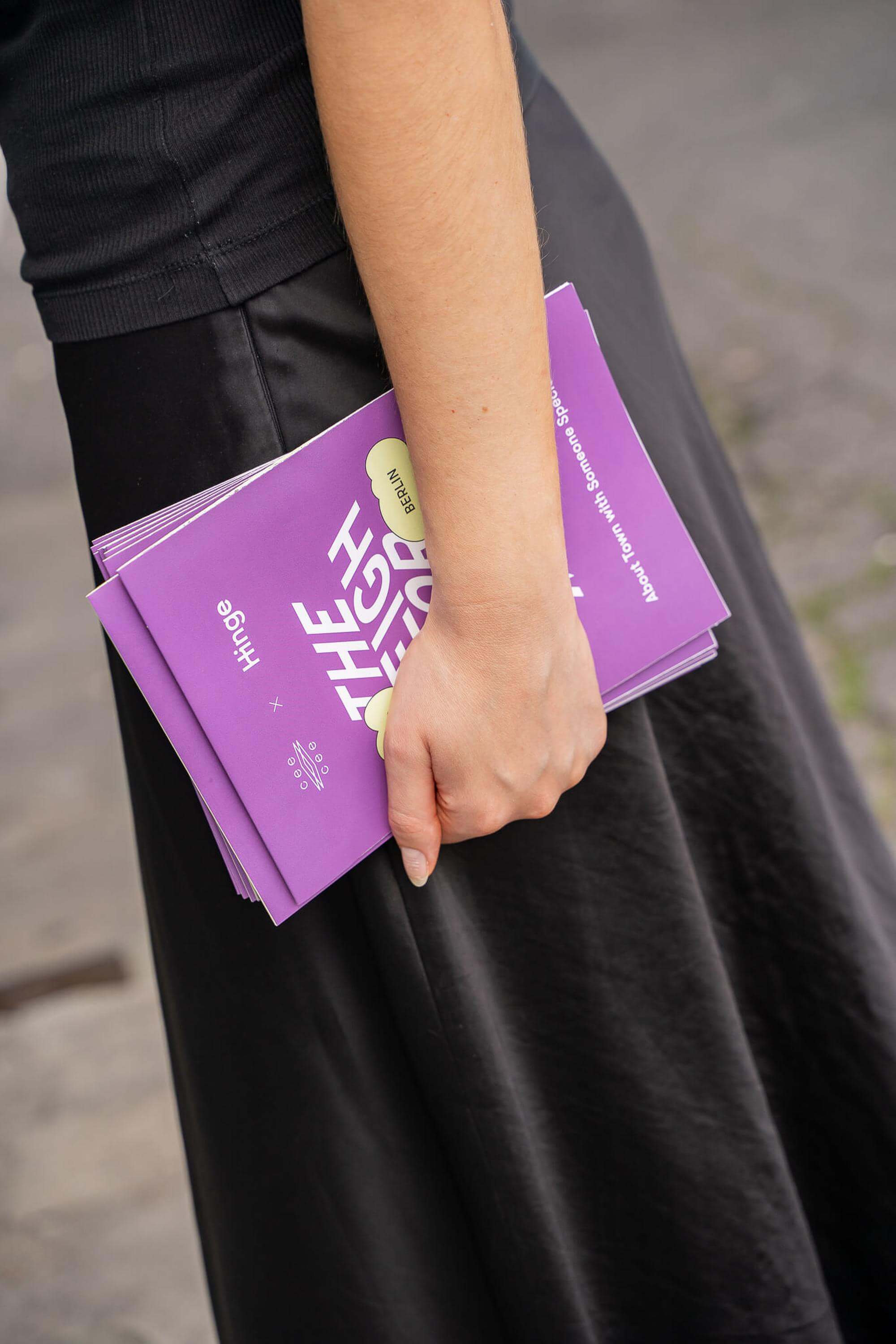 A person in a black outfit holds several purple “The Neighborhood Map” brochures from the Hinge × Cee Cee Creative collaboration.