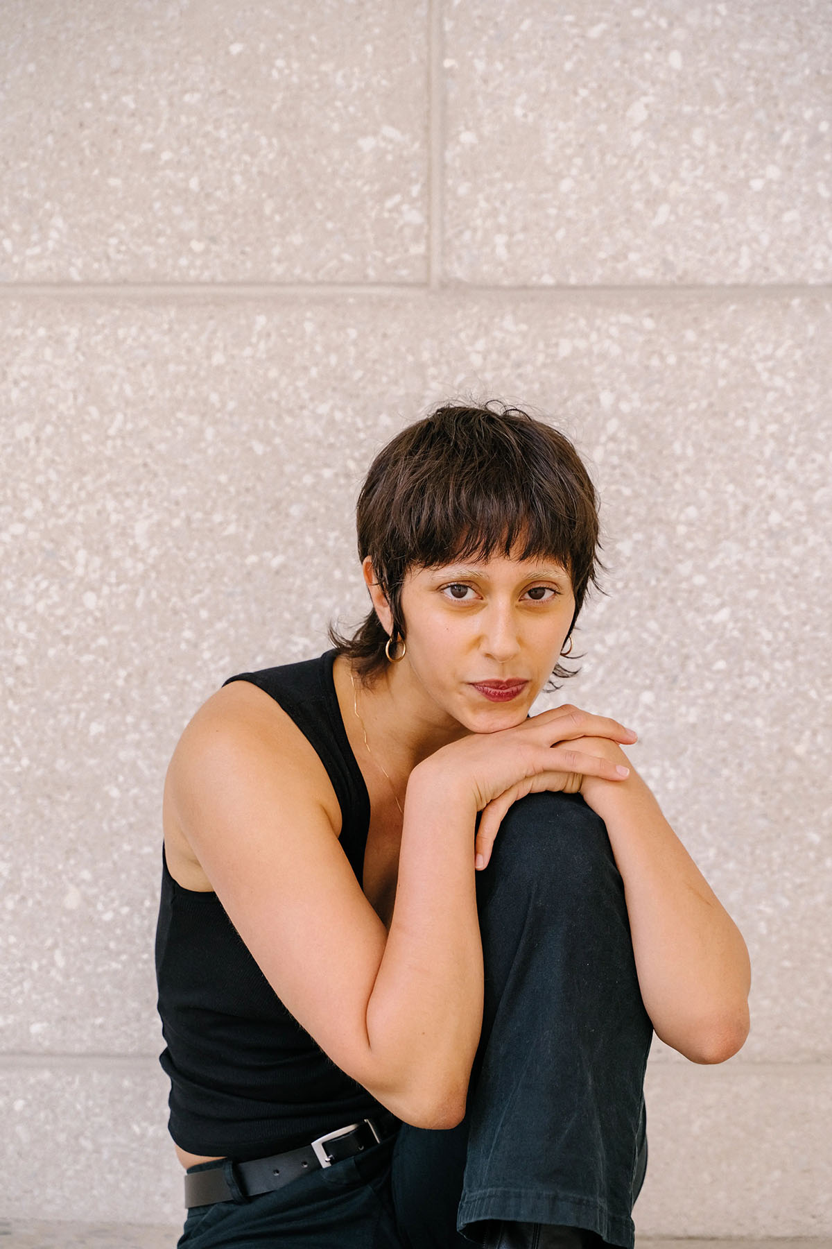 A person sits in front of a textured stone wall with their arms resting on their knee, looking directly into the camera.