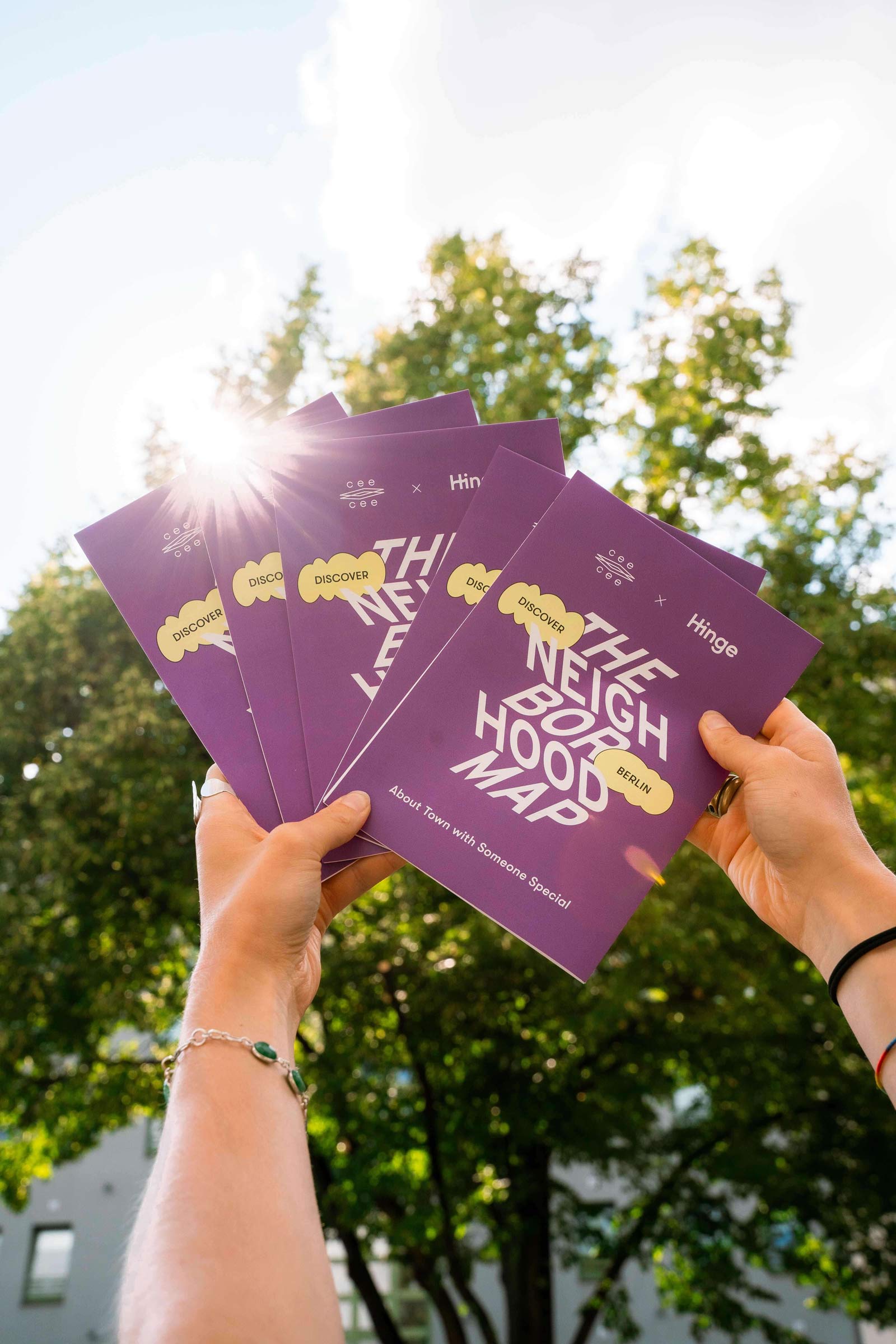 Multiple hands hold up several purple “The Neighborhood Map” brochures outdoors with sunlight shining through the trees.