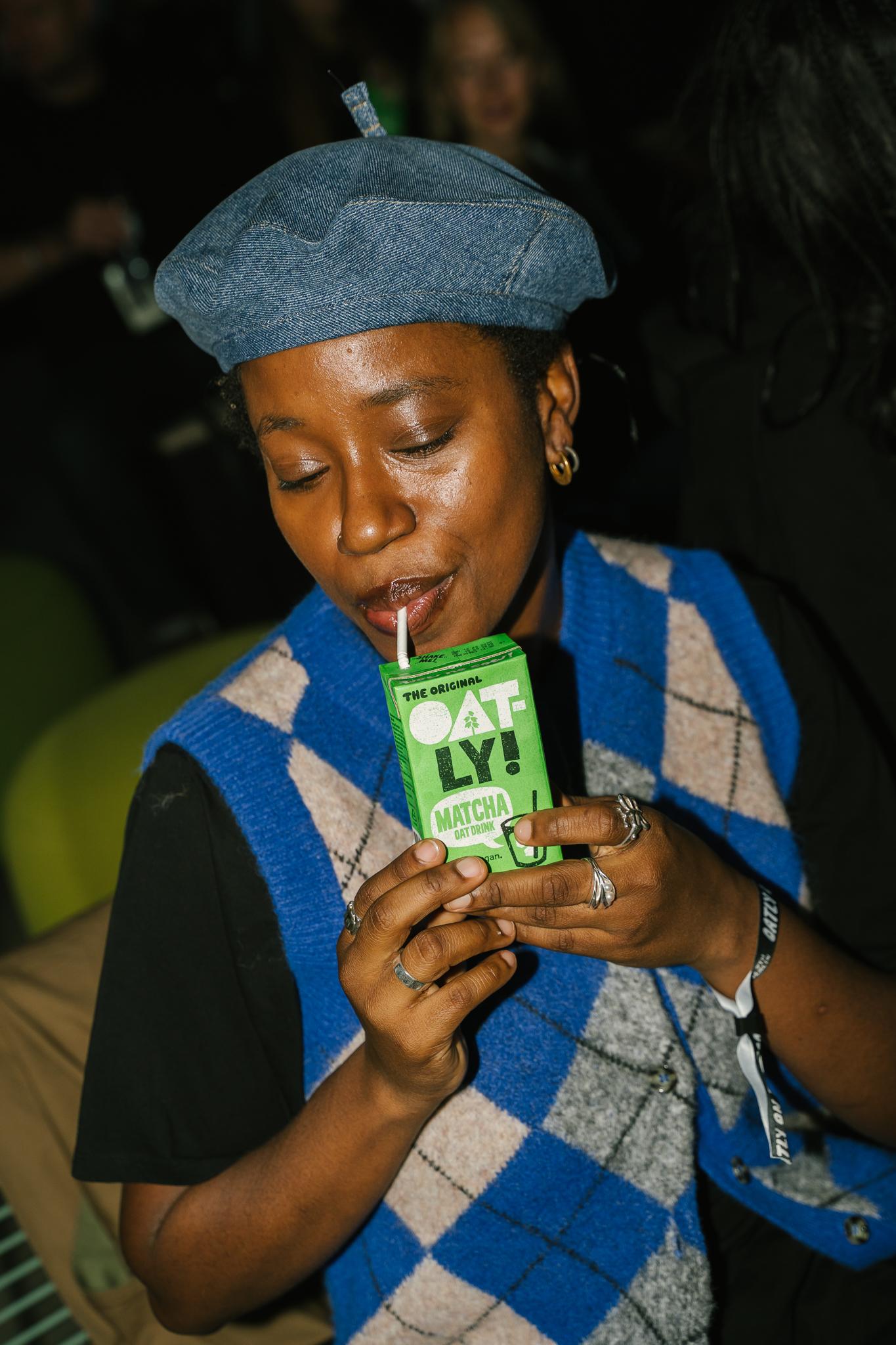 A guest sipping an Oatly Matcha drink through a straw, holding the green carton while smiling and wearing a blue-and-grey patterned vest and denim beret at the launch party.