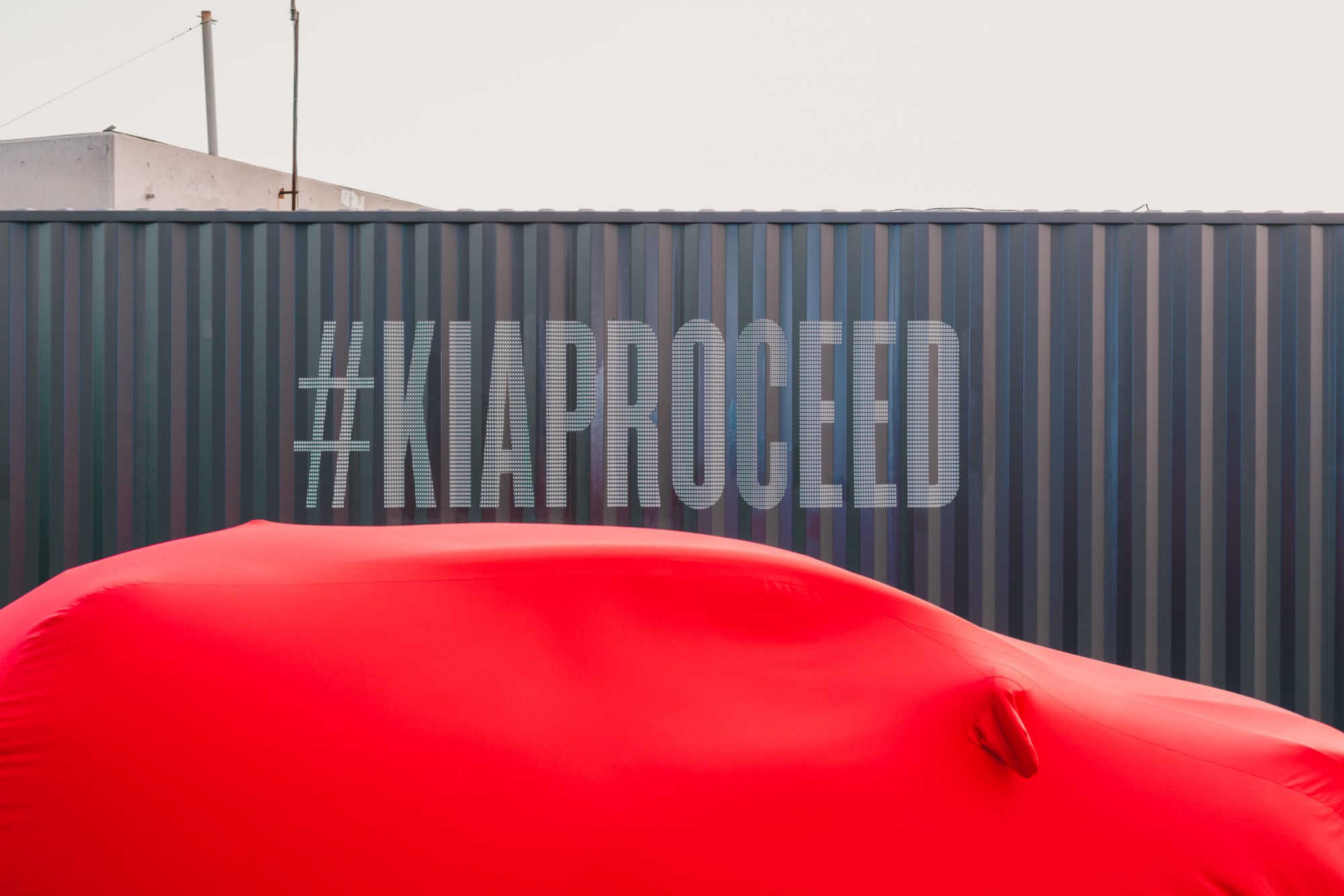 A photograph of the covered new KIA car at the KIA launch event with the key visual in the background.