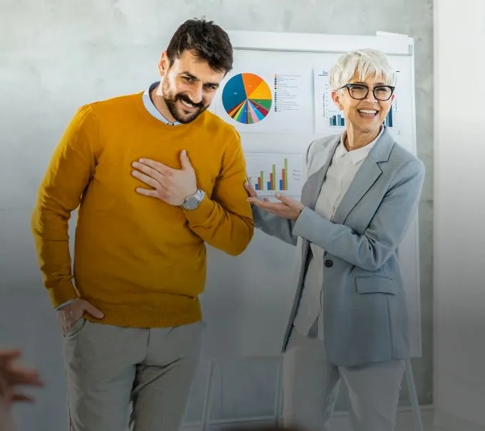Smiling older woman presenting a younger man who is laughing with hand on his chest, in front of a flipchart with charts.