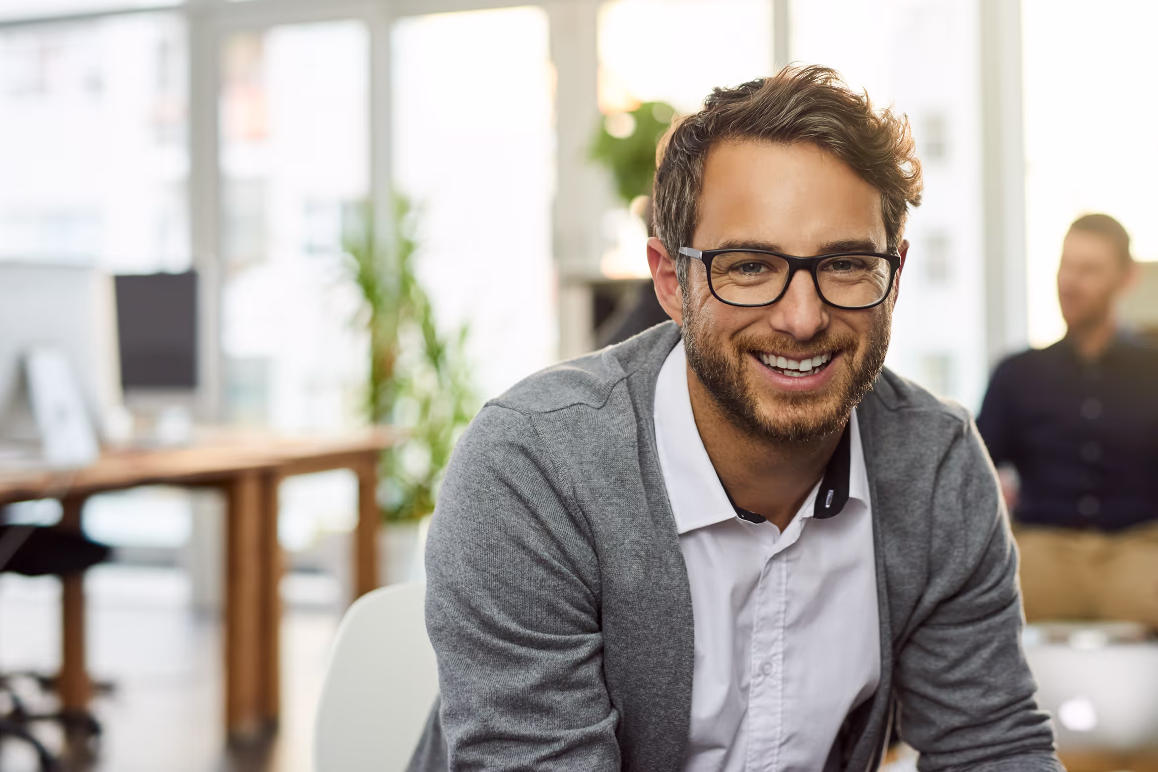 Bildet viser en smilende mann med briller og skjegg, iført en lys skjorte og en grå cardigan. Han sitter i et lyst og moderne kontorlokale med store vinduer og planter i bakgrunnen.