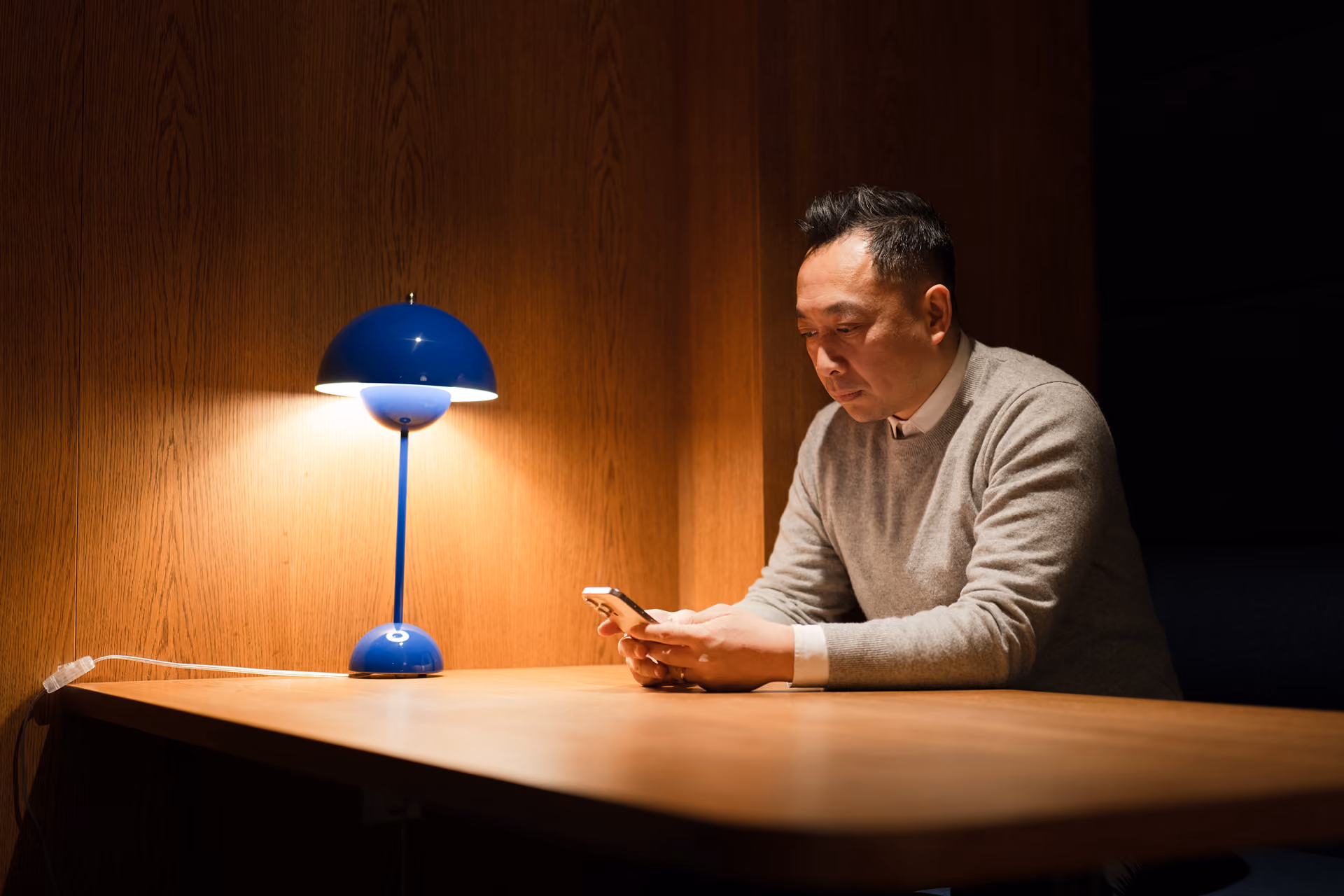 Mann sitter ved et bord i et svakt opplyst rom og ser på mobiltelefonen sin ved siden av en blå bordlampe