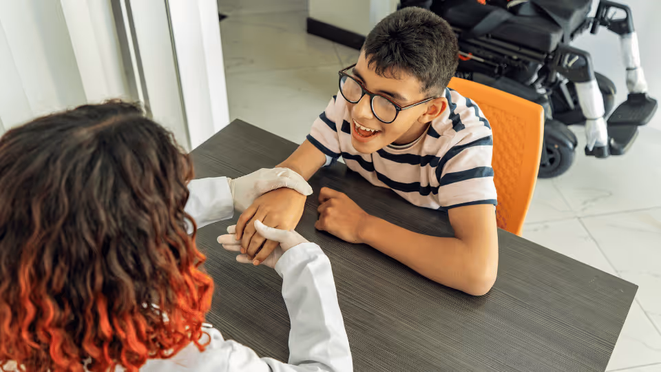 A boy wearing glasses and an occupational therapist are seated together at a table, engaged in conversation