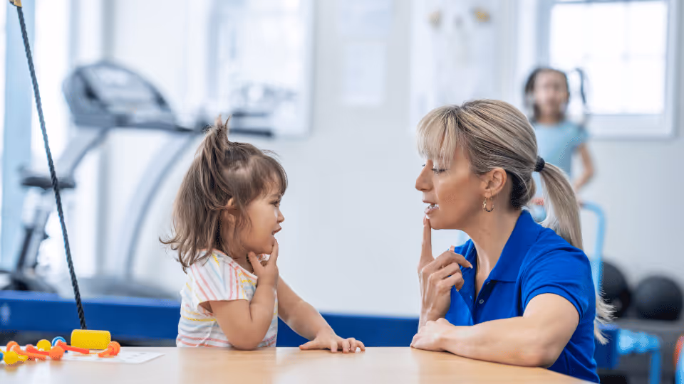 A woman teaching a child at a table in a gym, conducting a speech therapy session.