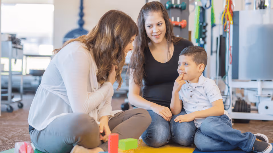 A therapist and two children are seated on the floor, surrounded by various toys, engaged in playtime together.