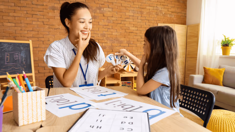 A woman and a child sit at a table covered with letters, as the occupational therapist guides the child's learning.