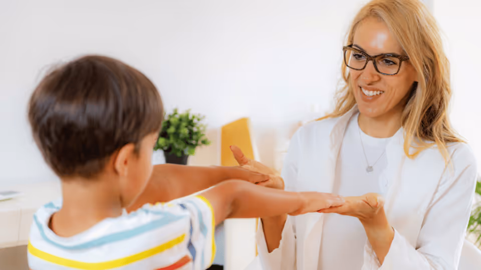 A woman holds a child's hand, both smiling, during a joyful moment in physical therapy with a therapist.