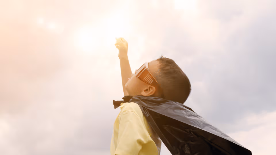A young boy wearing a cape and mask reaches upward, his arms extended towards the sun, symbolizing hope and dreams.