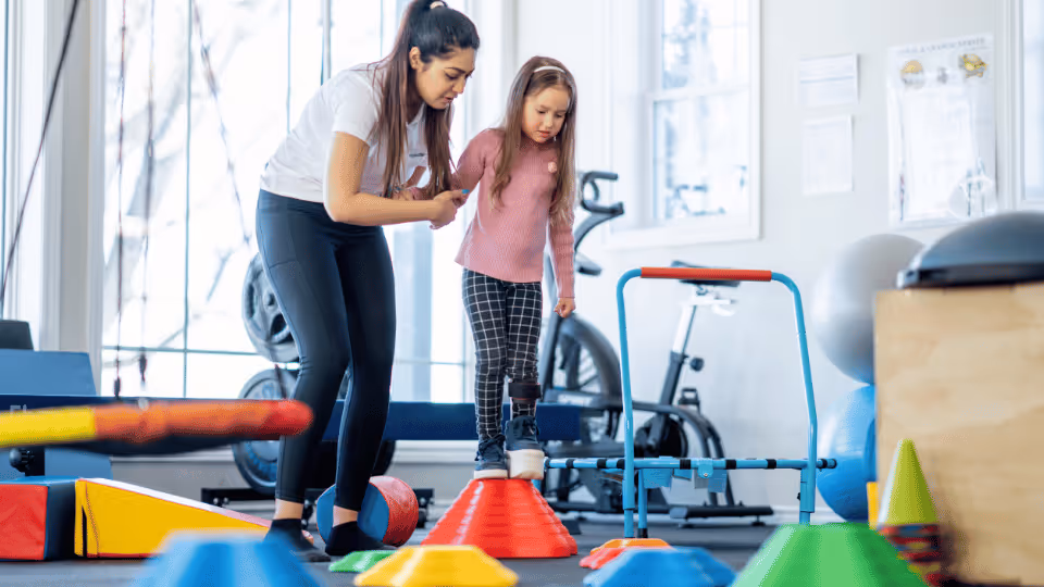 A physical therapist guides a woman and child in a gym, surrounded by colorful cones during a therapy session.