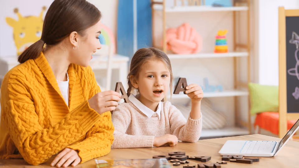 A woman and child engaged in a speech therapy session at a table, using a laptop for interactive learning.