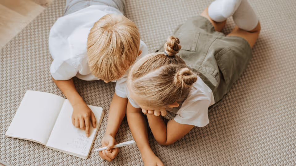 Two children engaged in occupational therapy, lying on the floor and writing in a book, fostering creativity and learning.
