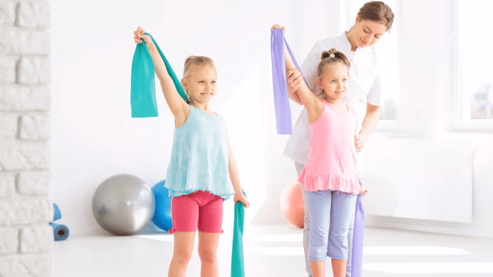 Two young girls engage in physical therapy exercises using resistance bands to enhance strength and flexibility.