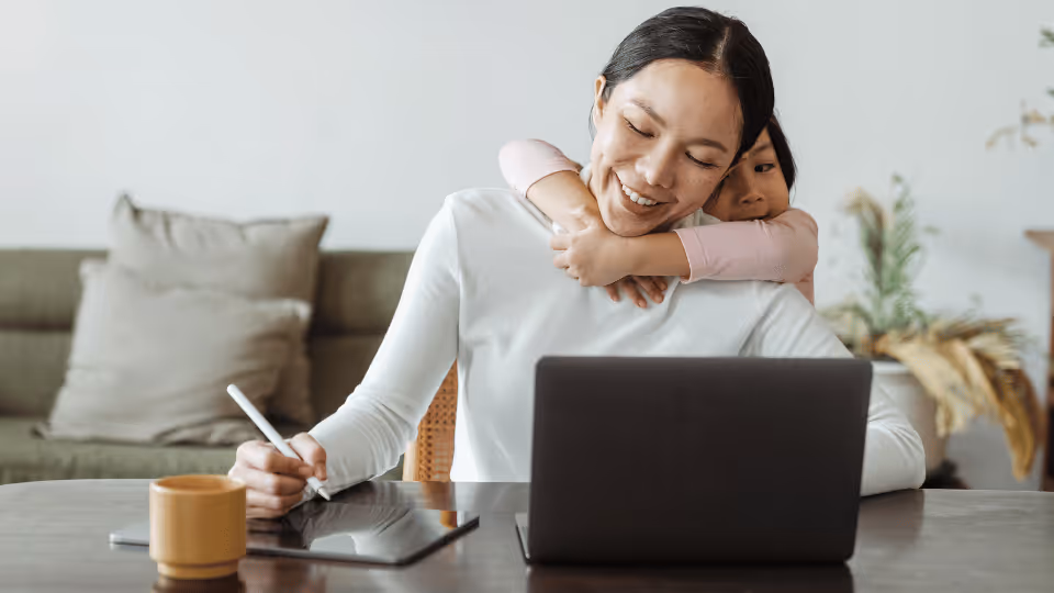 A woman sits at a table with her laptop, engaging with a child, exploring in-home pediatric physical therapy options.