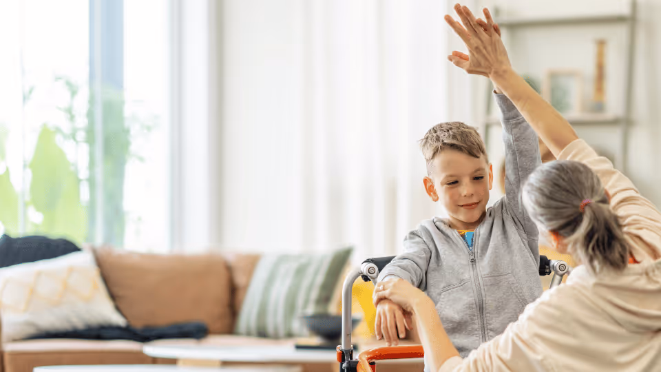 A woman and a boy in a wheelchair joyfully raise their hands during in-home pediatric physical therapy session.