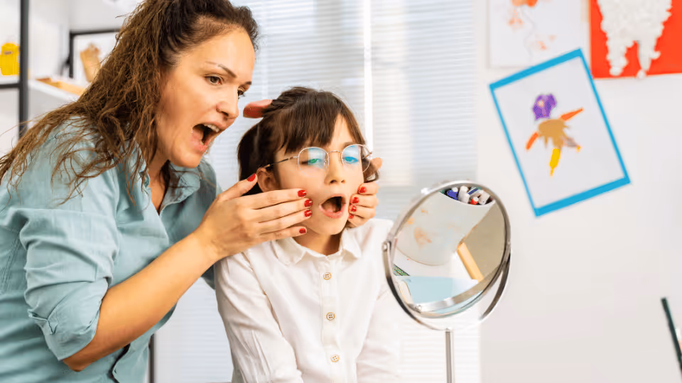 A woman and a child engage in pediatric therapy, observing their reflections in a mirror together.