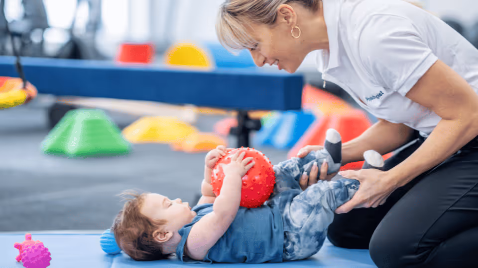 A woman assists a child in a gym, focusing on developing fine motor skills while playing with a ball.