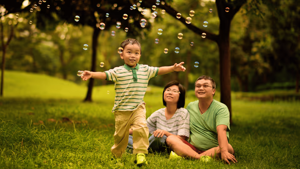 A family of three sits on the grass, engaging in activities to support gross motor skill development.